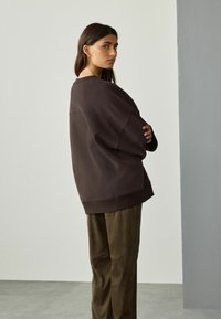 Frau mit langen, dunklen Haaren, die einen lockeren, dunkelbraunen Pullover und olivgrüne Hosen trägt, und über ihre Schulter gegen eine schlichte Wand schaut.