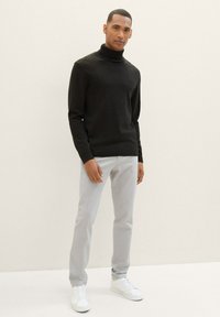 Zwarte coltrui in een soepele breistekstuur, gecombineerd met lichtgrijze chino's en witte sneakers. Model staat met de handen langs het lichaam.