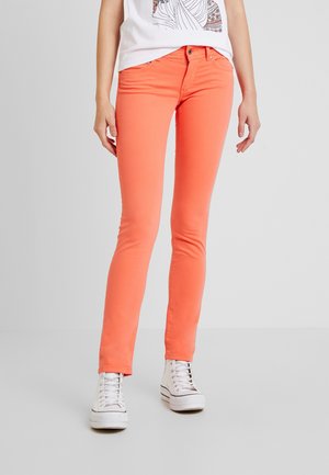 Femme portant un pantalon slim corail, un t-shirt blanc à motif et des baskets montantes blanches, debout devant un fond gris uni.