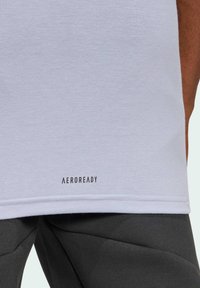 Camiseta gris claro hecha de una tela suave con corte recto, que presenta un logo negro de "AEROREADY" en el dobladillo inferior. Combinada con pantalones oscuros.