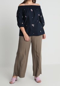 Femme portant un haut bleu marine dénudé d'épaules avec des broderies florales, un pantalon large taupe et des mules à talons ouverts roses, se tenant contre un fond uni.