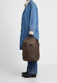 Sac en tissu marron avec une poignée, de forme rectangulaire, arborant une étiquette noire. Porté par une personne en manteau en denim et en jeans ajustés.