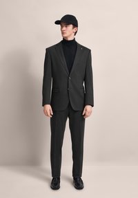 Dunkelgrauer Anzug mit zwei Knöpfen, passenden Hosen und einem schwarzen Rollkragenpullover. Akzentuiert mit einer schwarzen Mütze und schwarzen Schuhen, minimalistischem Design.