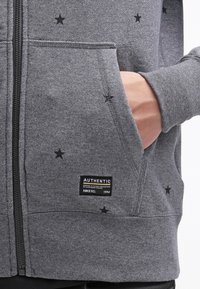 Szara bluza rozpinana z miękkiego materiału, ozdobiona małymi czarnymi gwiazdkami, z przednimi kieszeniami oraz metką z napisem „AUTHENTIC” i „NIKE FC 1994”.