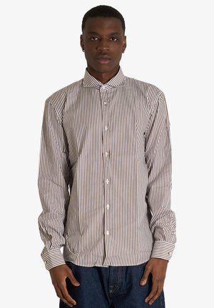 Giovane uomo con indosso una camicia a maniche lunghe a righe verticali marroni e bianche e jeans blu scuro, in piedi rivolto in avanti.