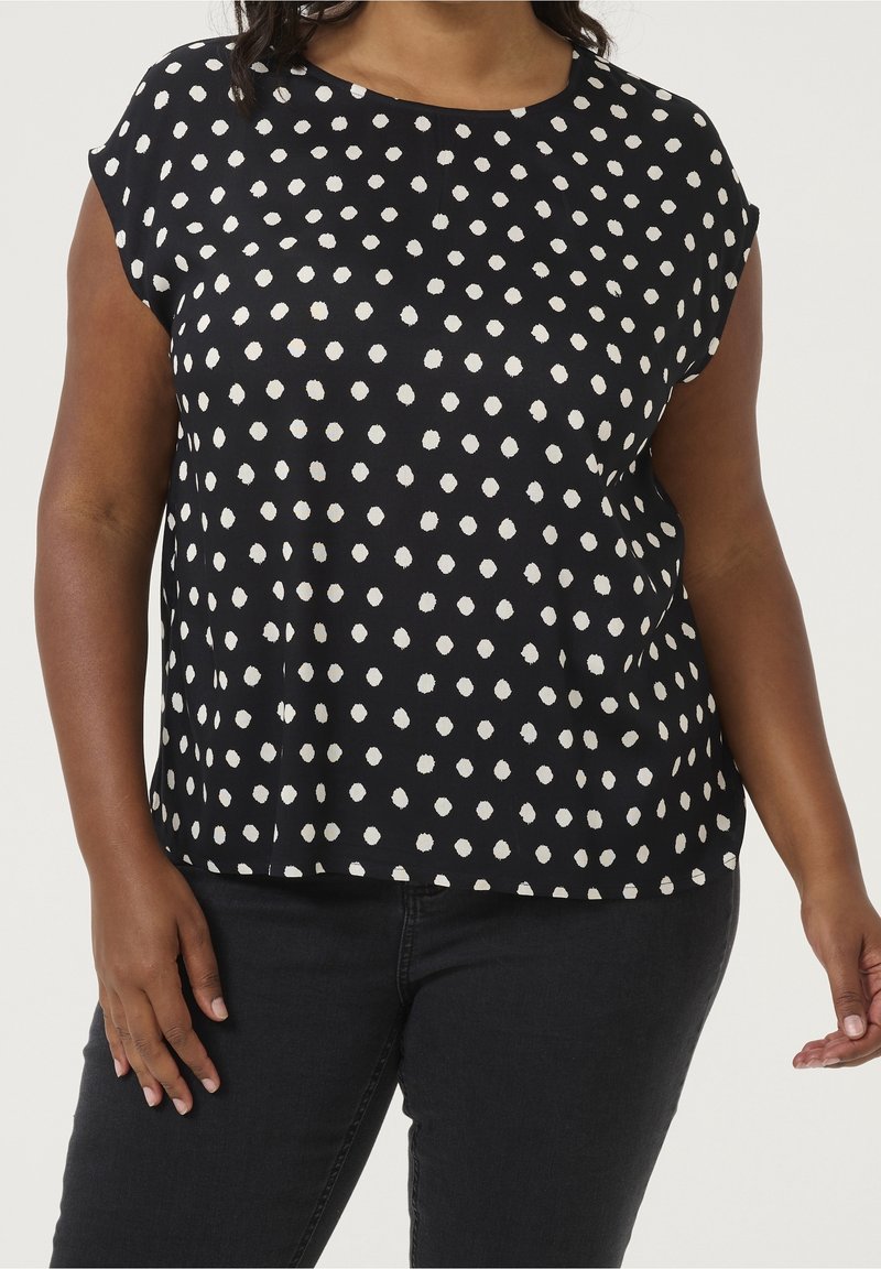Blouse noire avec motif à pois blancs, col rond, manches courtes et coupe ample. Tissu lisse avec fentes latérales à l'ourlet.