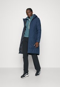 Marinblå pufferjacka med huva, med en frontdragkedja och två sidofickor, i kombination med en teal-fleece och svarta byxor. Sneakers fulländar outfiten.