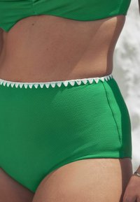 Bikini donji dio visokog struka od teksturirane zelene tkanine s bijelim valovitim obrubom duž struka. Glatka završna obrada, dizajn koji se prilagođava tijelu.