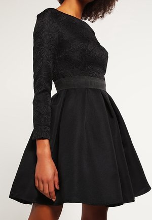 Robe noire à manches longues, texture fleurie sur le corsage, taille cintrée, jupe évasée et tissu lisse. Coupe et silhouette classiques.