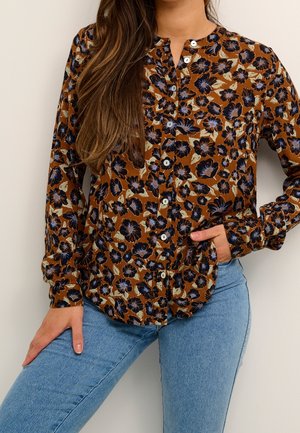 Kobieta w brązowej bluzce zapinanej na guziki z czarnym i beżowym kwiatowym wzorem, w połączeniu z jasnoniebieskimi dżinsami, pozująca z jedną ręką w kieszeni.