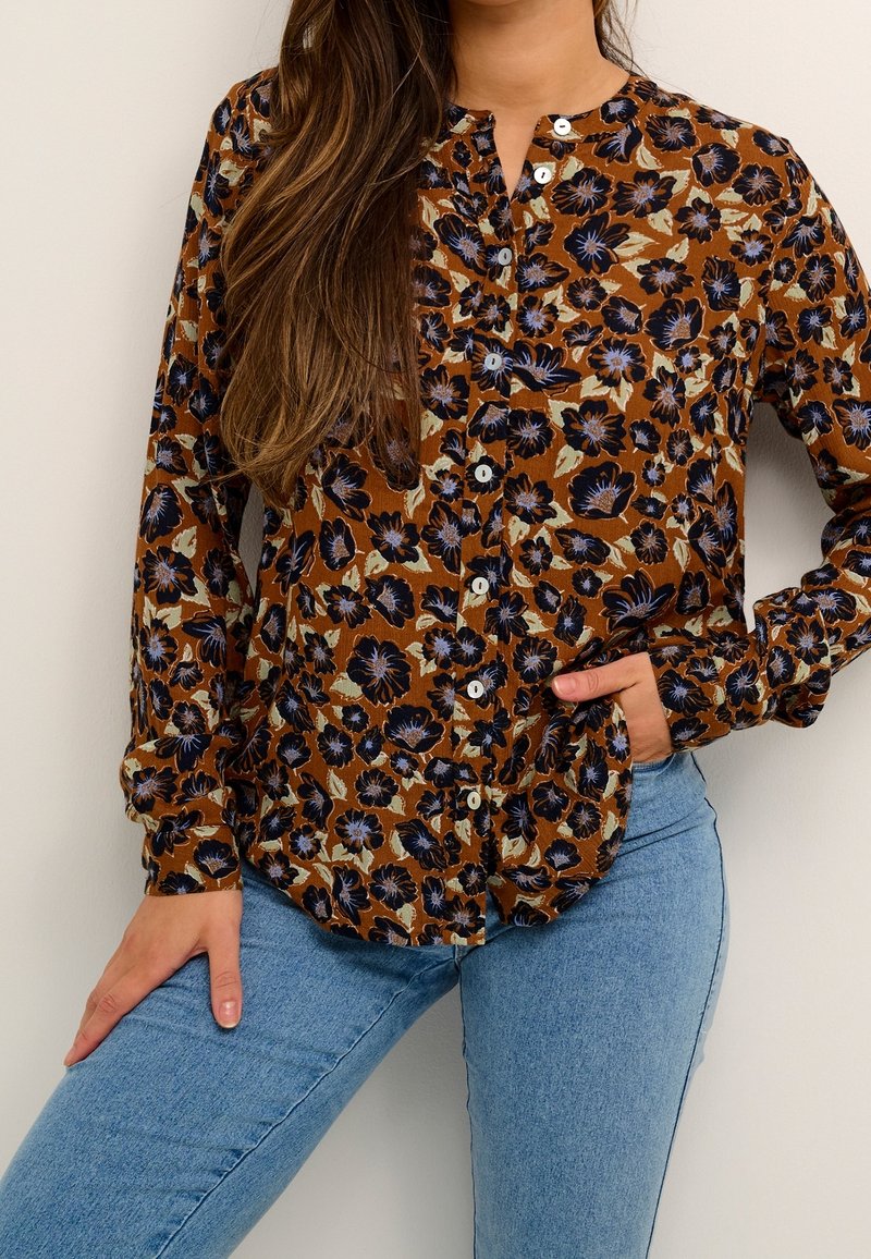 Femme portant un chemisier boutonné marron avec un imprimé floral noir et beige, associé à un jean bleu clair, posant avec une main dans la poche.