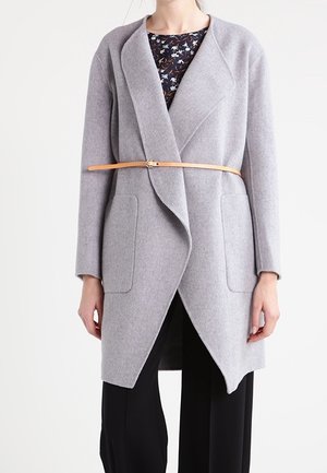 Femme portant un manteau enveloppant gris clair avec de grandes poches avant, ceinturé à la taille avec une fine ceinture beige, sur un pantalon noir et un top fleuri.