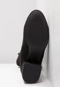 Czarny zamszowy botek na kostkę z teksturowaną gumową podeszwą, średnim szerokim obcasem i zamkiem błyskawicznym z boku. Podeszwa ma poziome rowki zwiększające przyczepność.