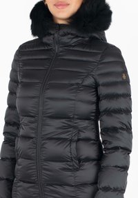 Giacca a vento nera con design trapuntato, chiusura con zip e cappuccio in pelliccia sintetica. Il materiale appare lucido e leggero.