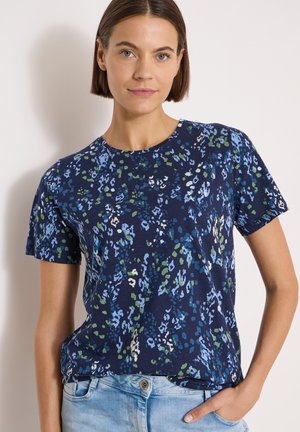 Femme aux cheveux courts et bruns portant une chemise à manches courtes à motif floral bleu marine et un jean bleu clair, une main dans la poche.