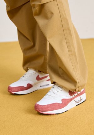 Person iført beige bukser og hvide sneakers med lyserøde ruskind accenter står på en gul gulv.
