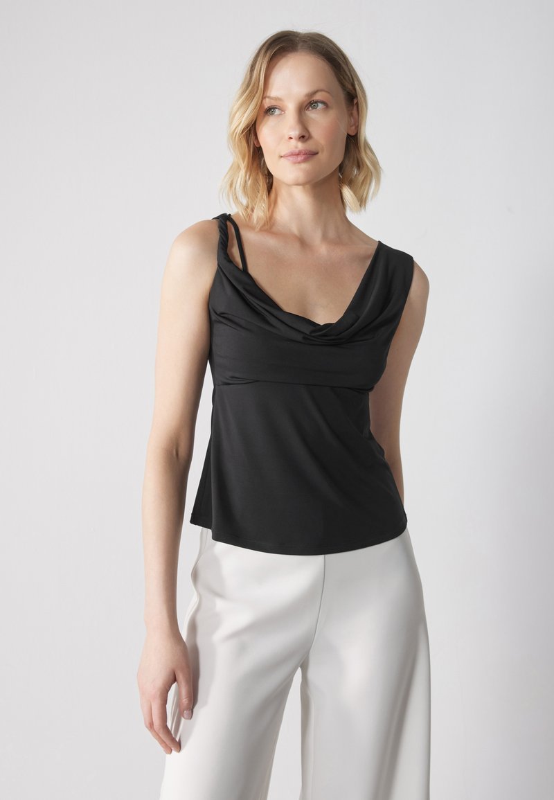Svart utan ärmar topp med en draperad halsringning, tillverkad av mjukt material, kombinerad med vita vida byxor som skapar en kontrasterande look.