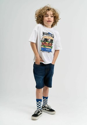 Biała koszulka z graficznym wzorem burgera, niebieskie szorty dżinsowe, czarne sneakersy i niebieskie skarpetki w kratkę. Kręcone włosy.