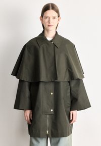 Casaco verde com gola, apresentando um design de capa em camadas, fecho frontal de correr, botões de pressão e dois bolsos laterais. Textura de tecido suave.