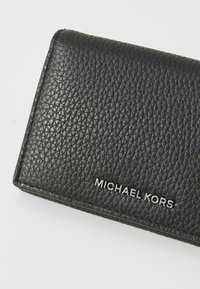 Zwarte leren portemonnee met een textuur oppervlak en een metallic "MICHAEL KORS" logo. Compacete rechthoekige vorm met strakke randen.