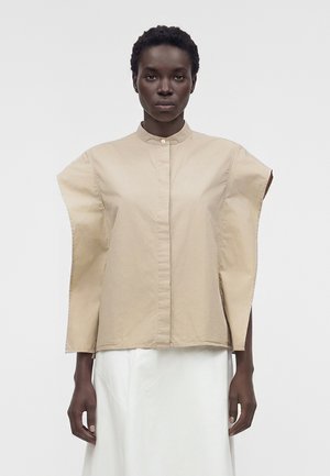 Kobieta w beżowej oversize bezrękawnej koszuli z wysokim kołnierzem i białej spódnicy, stojąca na tle jednolitego jasnego tła.