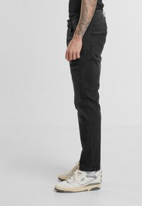 Jeans de denim negro mostrados de lado, con un corte slim, un leve desgastado y un diseño de cinco bolsillos. Combinados con zapatillas blancas.