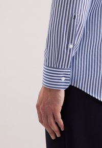 Camicia a righe blu e bianche con polsini abbottonati, caratterizzata da righe di larghezza variabile e un ricamo sottile sulla manica. Tessuto di cotone.