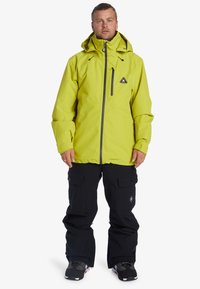 Bright green waterproof jacket with a hood, featuring a zip pocket, paired with black pants. The outfit is designed for winter activities.