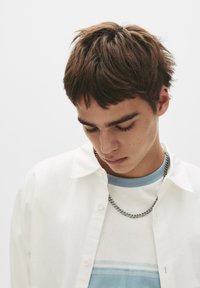 Weißes Hemd über einem hellblauen T-Shirt mit horizontalen Streifen, dazu eine Silberkette. Kurze, strukturierte braune Haare.