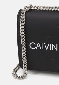 Černá crossbody taška z umělé kůže se stříbrným řetězovým popruhem. Obsahuje výrazné bílé logo "CALVIN" a hladký povrch s minimalistickým designem.