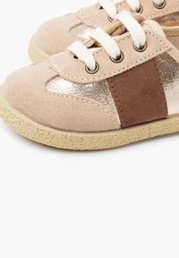 Béžové semišové boty s kovovými zlatými doplňky a hnědým semišovým panelem. Mají bílé tkaničky a texturovanou gumovou podrážku.