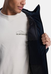 Marine isoleret jakke vist uaf zipper over en lys grå sweatshirt med et Quiksilver-logo. Blød tekstur, kontrasterende materialer og rene linjer.