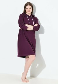 Junge Frau steht barfuß und trägt ein langärmeliges lila Kapuzenkleid, die Arme verschränkt, vor einer weißen Wand mit sanften Schatten.