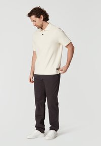 Kortärmad krämfärgad polo med texturerad tyg, med en standardkrage och en svart knapp. Kombinationen med mörka byxor och vita sneakers.