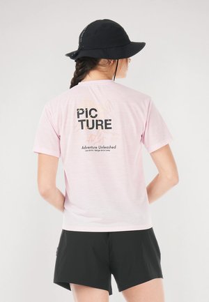 Svetloružové tričko s textom „PIC TURE“ a grafikou hôr na chrbte, doplnené o čierne šortky a čierny klobúk typu bucket.