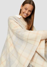 Poncho à carreaux crème et gris avec un motif à damier orange, fabriqué dans un tissu doux. Présente un bord frangé et une coupe ample.