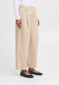 Pantaloni beige a gamba larga con pieghe frontali, realizzati in tessuto leggero; abbinati a scarpe nere basse con fascia e piccoli dettagli in metallo.