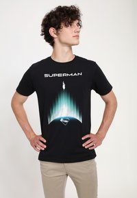 Czarna bawełniana koszulka z nadrukiem logo Supermana oraz grafiką postaci wznoszącej się w górę, z niebieskimi i zielonymi akcentami w tle.