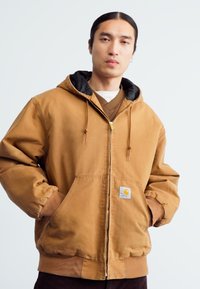 Veste à capuche brune zippée avec doublure noire, poches avant et poignets élastiques. Présente une patch logo sur la poche gauche.