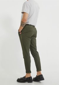 Olivově zelené kalhoty se zúženými nohavicemi, bočními kapsami, ohrnutými lemy a zadními kapsami, spojené s černými mokasíny. Bavlněná směs tkaniny.