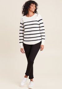 Gestreifter, weißer und marineblauer Strickpullover mit einem Rundhals-Ausschnitt, Knopfdetails an der Schulter und gerippten Bündchen, kombiniert mit schwarzen Jeans und weißen Sneakers.