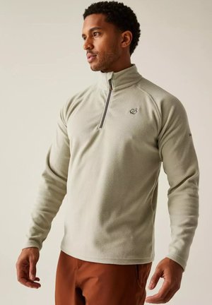 Hombre con un forro polar gris claro con cremallera hasta un cuarto y pantalones de color óxido, mirando a su izquierda.