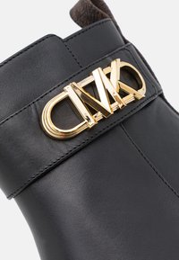 Botte en cuir noir avec boucle décorative métallique dorée affichant des lettres stylisées sur la tige supérieure.