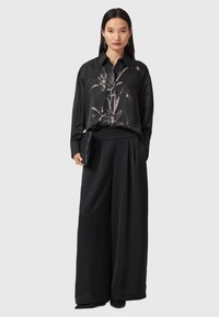 Femme portant un chemisier en soie noir à motif floral, un pantalon large noir, tenant une pochette noire, se tenant devant un fond clair. 