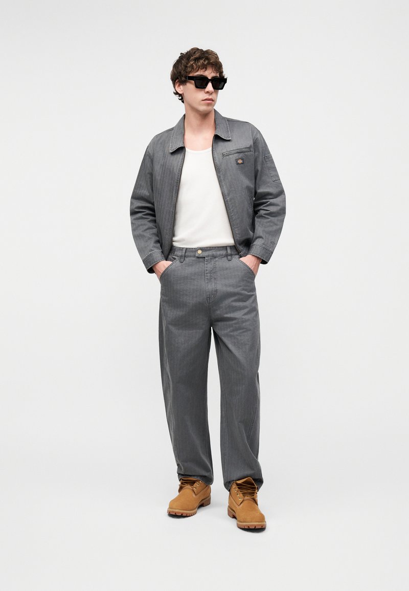 Jonge man met een grijze jas en broek, wit hemdje, beige werkschoenen en zwarte zonnebril, staand met handen in de zakken.