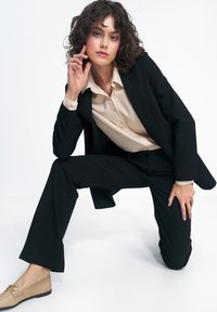 Kobieta z kręconymi włosami, ubrana w czarny garnitur i beżową koszulę, klęczy, opierając jedną rękę na nodze, a drugą blisko twarzy, na białym tle.