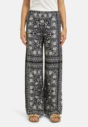 Woman wearing black wide-leg pants with white floral paisley patterns and beige sandals, standing against a plain background.