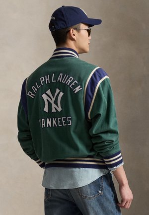 Grøn varsityjakke med "RALPH LAUREN" og "YANKEES" broderet på ryggen, med blå accenter og ribbede manchetter. Båret over en denimskjorte.