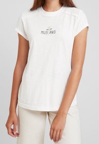 Camiseta blanca de manga corta con cuello redondo, que presenta bordado "MUSTANG" y un diseño de rosa en el frente. Tela suave y ligera.