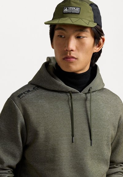 Sudadera verde texturizada con capucha ajustable, que presenta un logo en el hombro. El modelo también lleva una gorra bicolor con un logo.